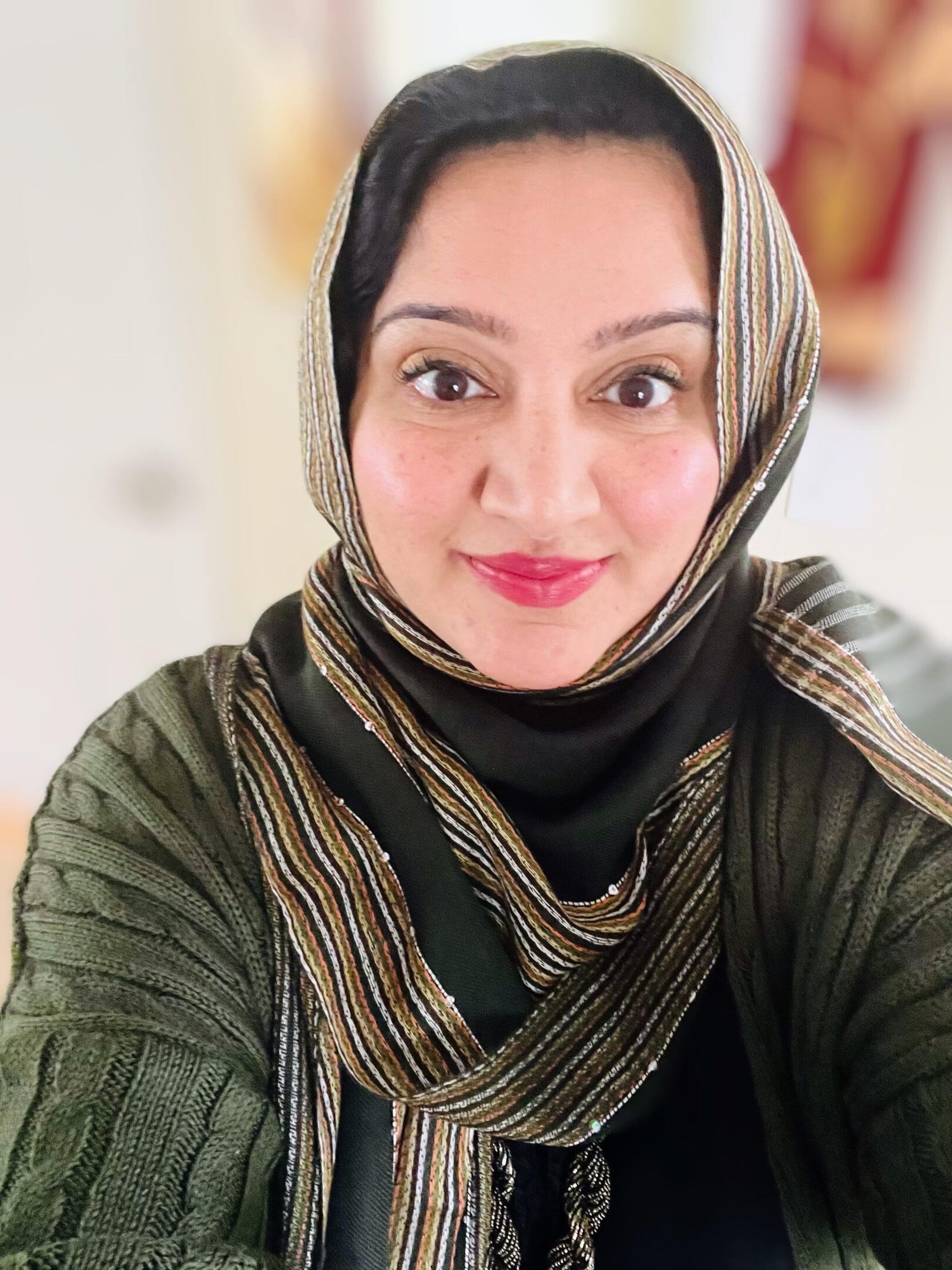 A woman wearing a striped hijab and green sweater smiling at the camera.